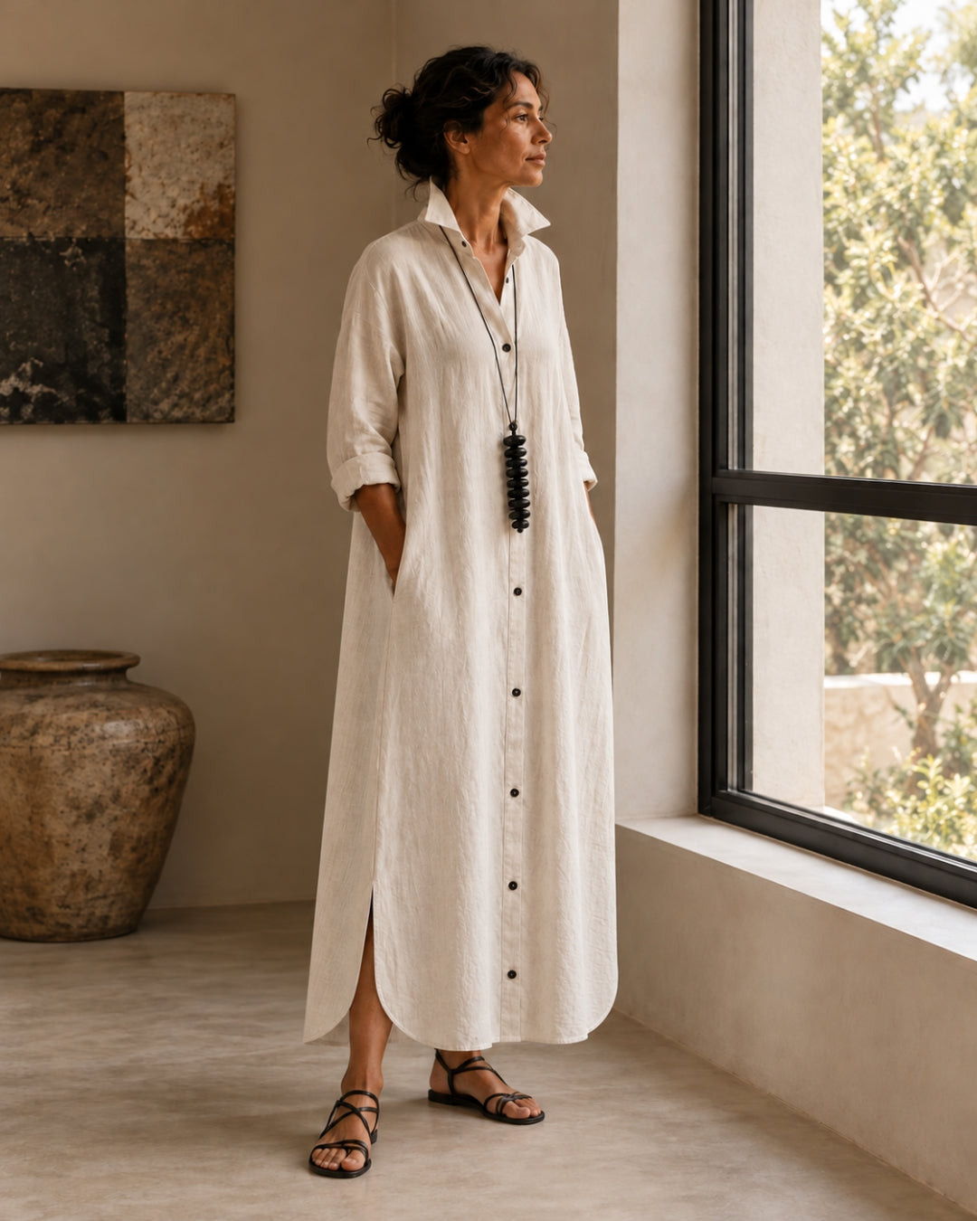 Full-length view of a model wearing the Jen Kahn Long Linen Shirt Dress in Natural beside a window, styled with a black pendant and flat sandals.