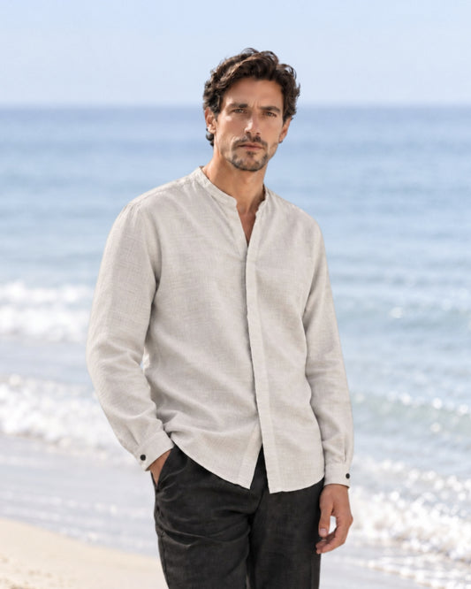Man standing on a beach wearing a jen kahn mandarin collar linen shirt linen long sleeve button-up shirt and dark trousers, with the ocean in the background.